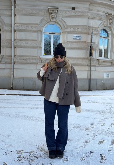 Abrigo corto marrón con detalle de lazo, suéter blanco de punto de cable, jeans azules, gorro negro y gafas de sol, de pie sobre un suelo cubierto de nieve.