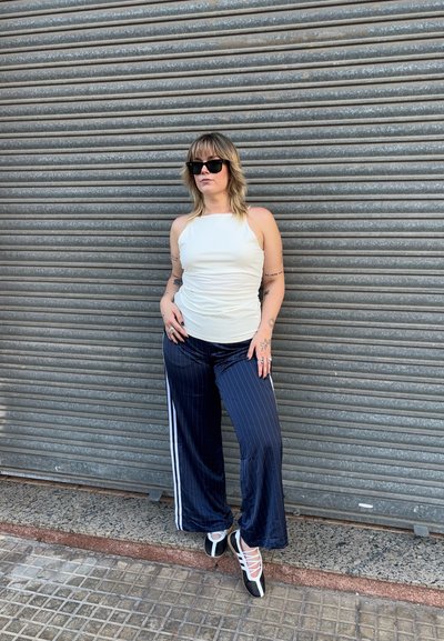 Una mujer está de pie contra una pared de metal corrugado, vistiendo una camiseta blanca ajustada, pantalones de pierna ancha a rayas marino con acentos blancos y zapatos negros.