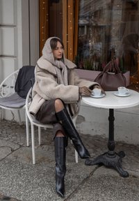 Manteau oversize en peau lainée beige, écharpe grise, jupe en cuir marron et bottes noires pointues jusqu'aux genoux. Table blanche avec tasses à café et sac à main.