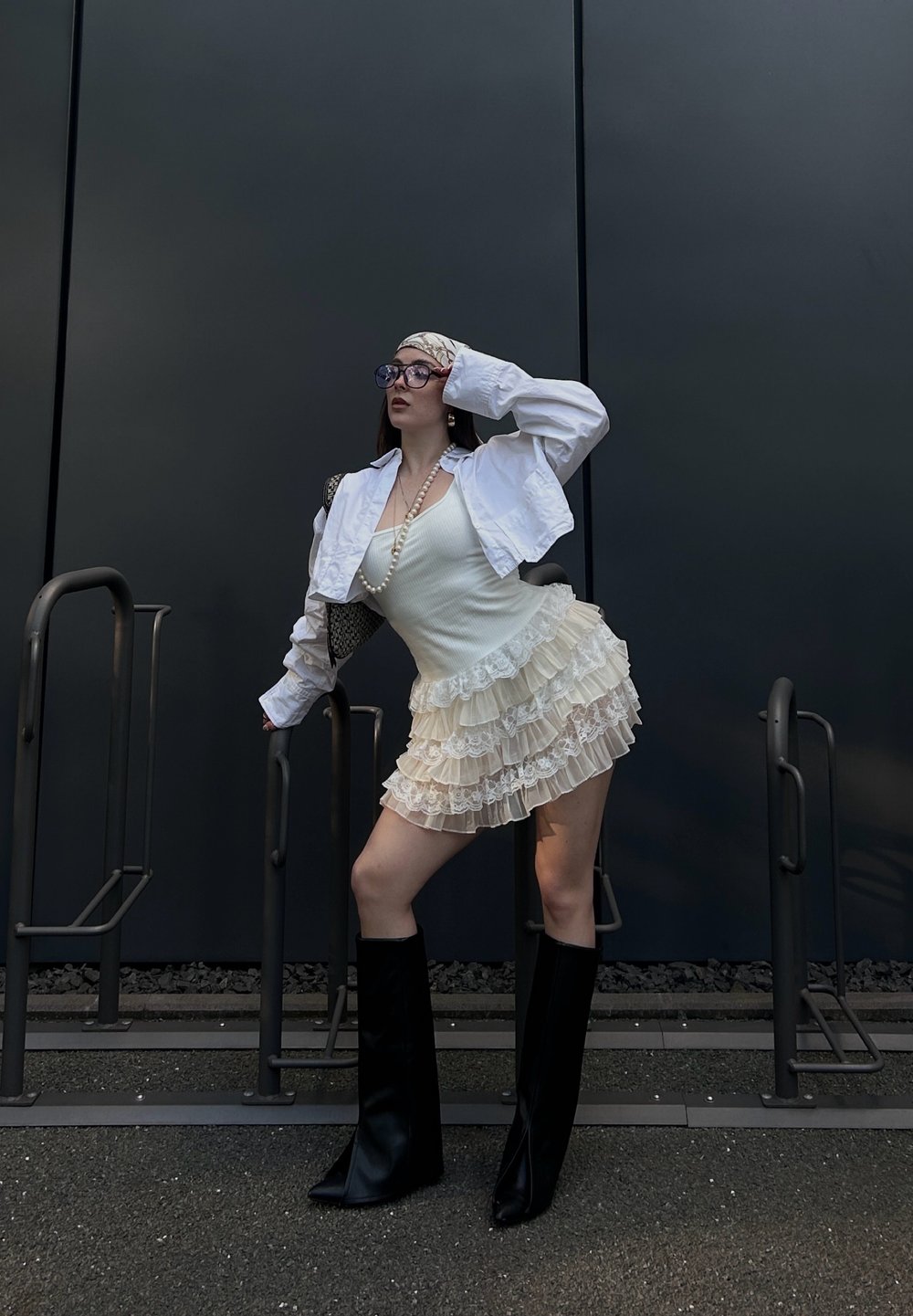 Jaqueta branca curta sobre uma blusa sem mangas creme e uma saia mini em camadas com babados. Botas pretas até ao joelho. Acessórios adicionais incluem óculos grandes e pérolas.