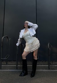 Jaqueta branca curta sobre uma blusa sem mangas creme e uma saia mini em camadas com babados. Botas pretas até ao joelho. Acessórios adicionais incluem óculos grandes e pérolas.