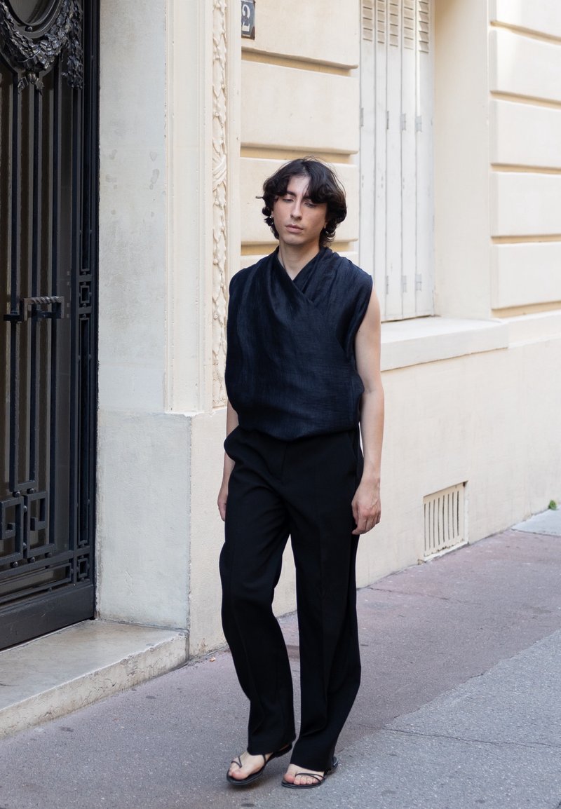 Jeune personne avec des cheveux ondulés porte un top noir sans manches, un pantalon noir et des sandales tout en se tenant sur un trottoir en ville près d'un bâtiment beige.
