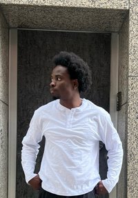 Camisa henley branca de manga longa com textura, com dois botões no pescoço, utilizada com calças pretas; em pé contra um fundo escuro texturizado.