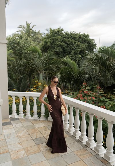 Vestido de sirena de punto marrón chocolate con un escote en V profundo, adornado con un broche dorado. La modelo sostiene un clutch de mimbre, de pie en un balcón de baldosas.