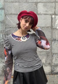 Pruhované tričko s dlhým rukávom a priehľadnými vzorovanými rukávmi, čierna sukňa a červený filcový baret; doplnené náhrdelníkom so špirálovým príveskom.