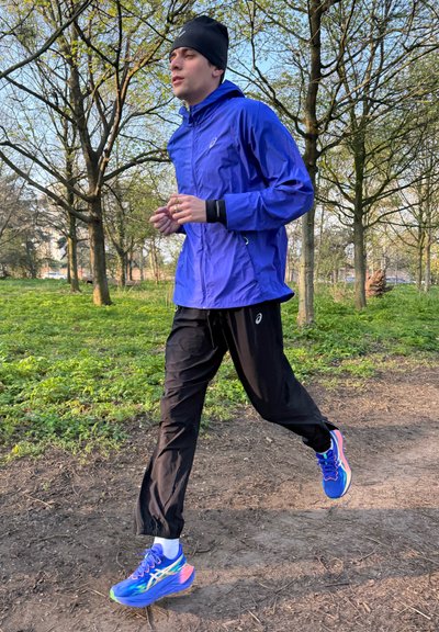 Joven corriendo por un sendero boscoso con chaqueta azul, pantalones negros, gorro negro y zapatillas azules para correr.