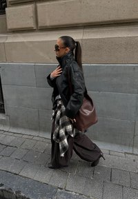 Sort læderjakke, oversized brune bukser, ternet flannel bundet om livet, brun håndtaske og solbriller; gående på en stensat sti.