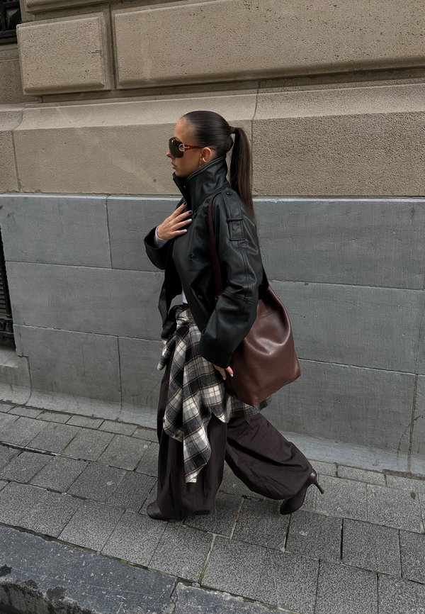 Sort læderjakke, oversized brune bukser, ternet flannel bundet om livet, brun håndtaske og solbriller; gående på en stensat sti.