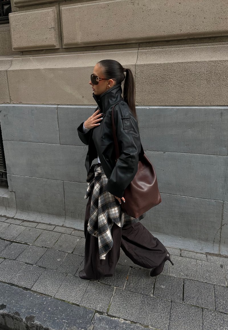 Veste en cuir noire, pantalon marron surdimensionné, chemise en flanelle à carreaux attachée à la taille, sac à main marron et lunettes de soleil ; marchant sur un chemin de pierre.