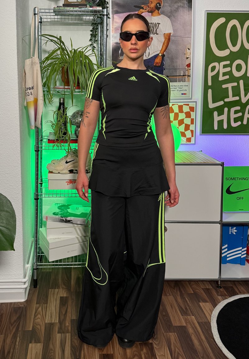 Black athletic set with short sleeves, featuring neon green stripes. Loose-fitting pants with a layered skirt overlay. Smooth texture. Sunglasses.