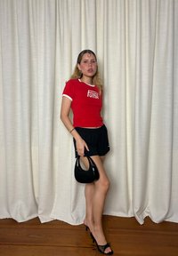 Ung kvinna i röd Puma-t-shirt och svarta shorts håller en svart handväska, bär svarta högklackade sandaler, står inomhus framför krämfärgade gardiner.
