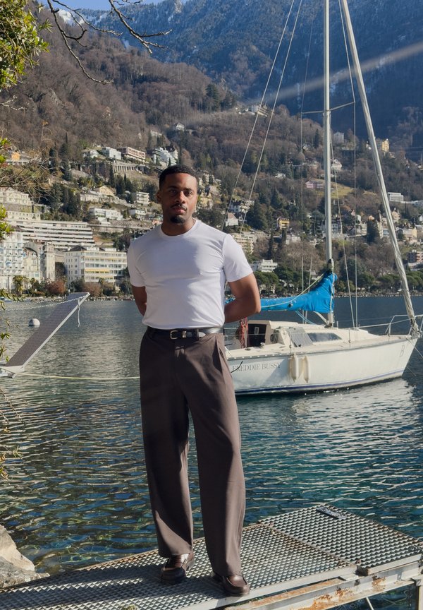 Homme en t-shirt blanc et pantalon foncé se tenant sur un quai métallique au bord du lac, avec un voilier et un village sur la colline en arrière-plan.