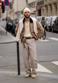 Tan Bomberjacke mit schwarzem Kragen, cremefarbenem Strickpullover, beigen Hosen und hellen Schuhen. Accessoirisiert mit einer goldenen Halskette und einem Schal.