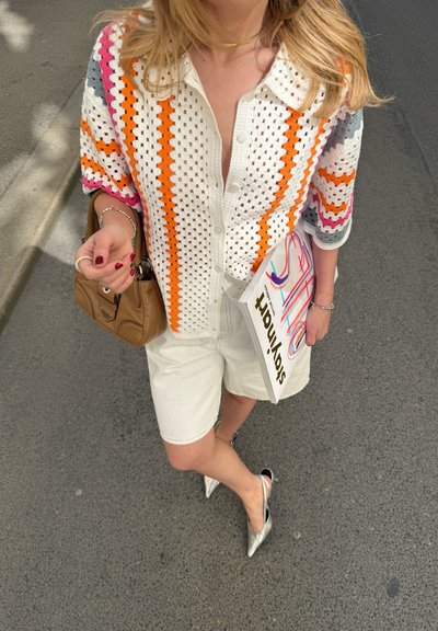 Top de punto multicolor con patrones en naranja, rosa y blanco, combinado con pantalones cortos crema y zapatos slingback plateados, sosteniendo una revista.