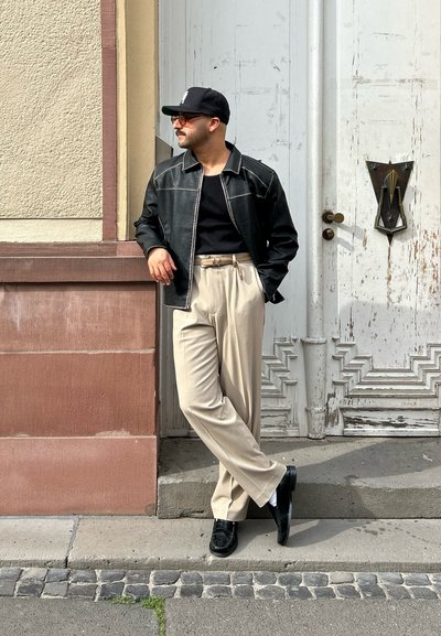 Chaqueta de cuero negra con costuras blancas, camiseta negra, pantalones de talle alto beiges y zapatos negros contra una pared texturizada.