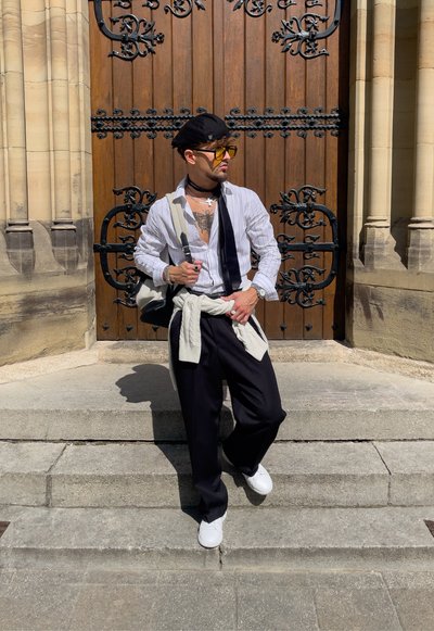 Pantalones de pierna ancha negros, zapatillas blancas, camisa a rayas y una boina negra. Los accesorios incluyen múltiples collares y un bolso llevado cruzado.