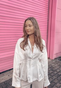 Blouse en satin blanc avec boutons, manches longues et coupe ajustée. En arrière-plan, un mur rose vif avec des lignes horizontales.