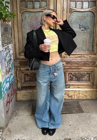 Sort cropped blazer, neongul cropped top med pink tekst, brede lysvaskede jeans, kraftige sorte sko, holder en kaffekop.