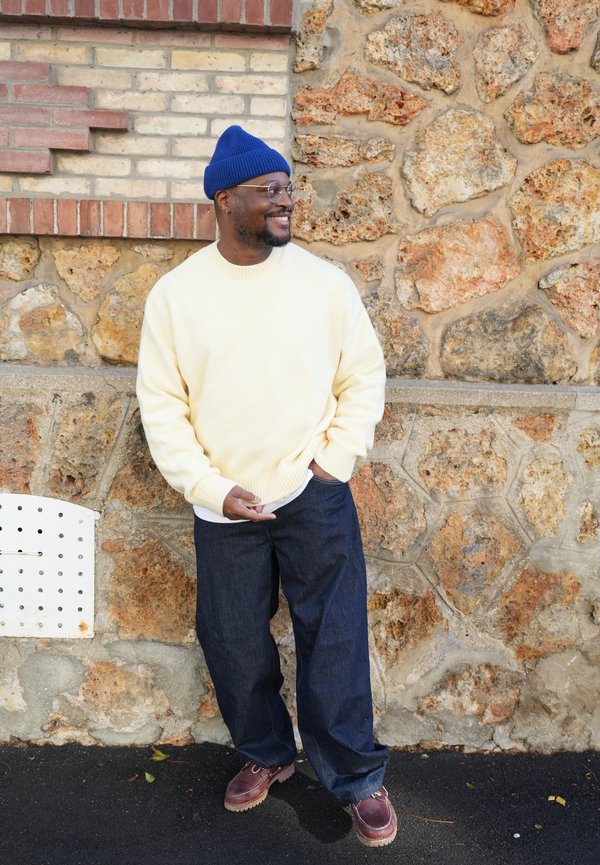 Un homme souriant, coiffé d’un bonnet bleu en maille et portant des lunettes, se tient dehors près d’un mur en pierre. Il porte un pull crème et un jean ample.