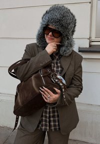 Sac en daim marron avec garniture argentée, porté sur une chemise à carreaux, associé à un chapeau gris en fourrure et de grandes lunettes de soleil noires surdimensionnées. Costume marron sur mesure.