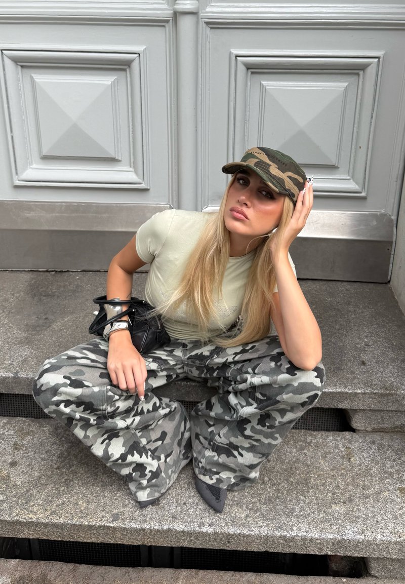 Housut leveällä lahkeella, harmaasävyisessä kamuflaasikuosissa, yhdistettynä kapeasuun vaaleanvihreään paitaansa ja kamuflaasi-lippikseen. Musta käsilaukku hopeisilla yksityiskohdilla.