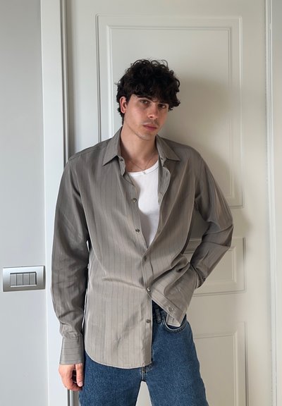 Camisa de manga larga de rayas gris claro con diseño de botones, llevada sobre una camiseta blanca de tirantes, combinada con jeans azules. Tejido suave, corte relajado.