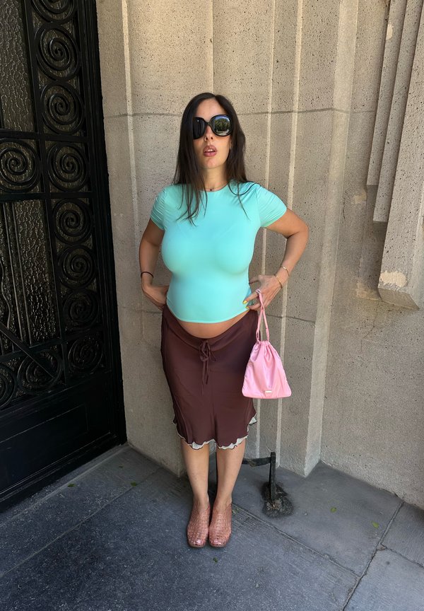 Femme en haut turquoise et jupe marron, debout contre un mur en pierre, portant de grandes lunettes de soleil noires et tenant un petit sac à main rose.