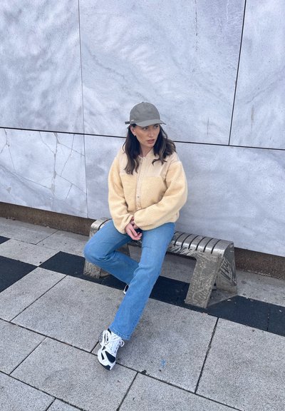 Mujer con chaqueta beige, vaqueros azules, zapatillas blancas y gorra gris sentada en un banco de metal frente a una pared de azulejos.