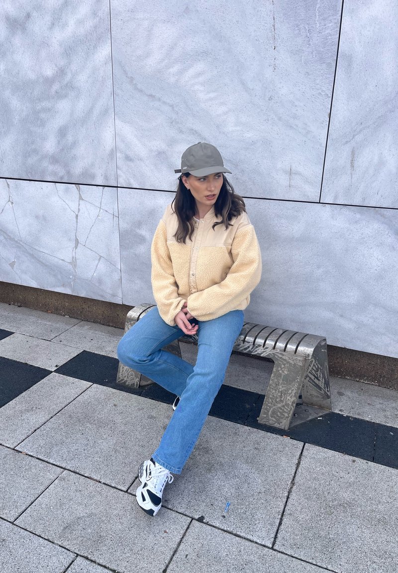 Frau in beige Jacke, blauen Jeans, weißen Turnschuhen und grauer Mütze sitzt auf einer Metallbank vor einer gefliesten Wand.