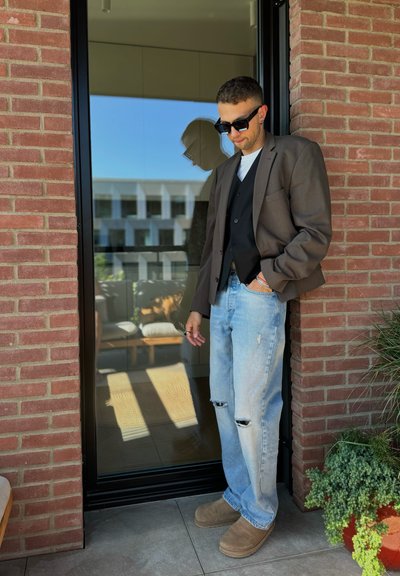 Blazer marrón sobre un chaleco negro, camisa blanca y jeans rasgados de color azul claro. Zapatillas de ante marrón. El fondo incluye una pared de ladrillo y un reflejo.