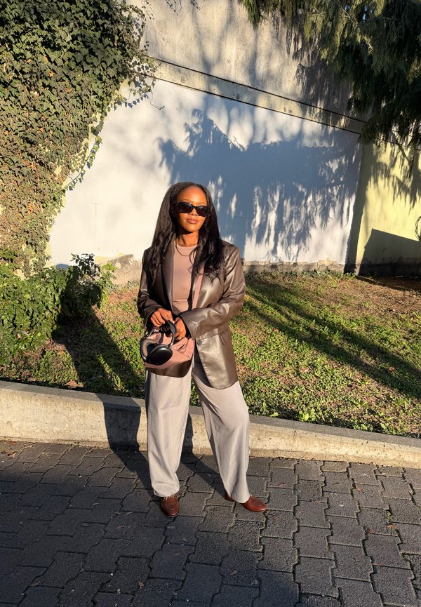 Femme portant des lunettes de soleil et une veste en cuir marron, debout sur un chemin pavé tenant un casque audio, avec des murs couverts de lierre et ensoleillés en arrière-plan.