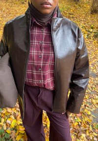 Veste en faux cuir marron avec un col haut portée sur une chemise à carreaux bordeaux. Associée à un pantalon marron, avec une texture lisse et une finition brillante.