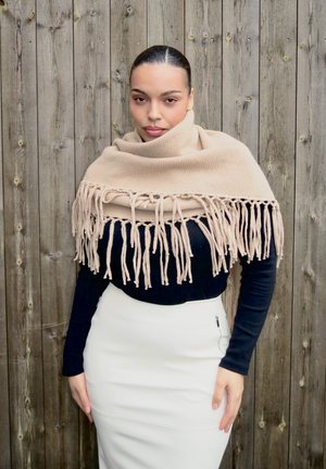 Mujer con el cabello oscuro recogido, llevando un chal beige con flecos sobre una blusa negra y una falda blanca, parada frente a una cerca de madera.