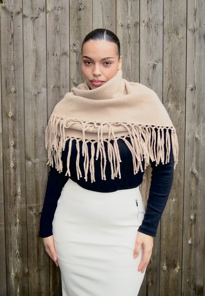 Mujer con el cabello oscuro recogido, llevando un chal beige con flecos sobre una blusa negra y una falda blanca, parada frente a una cerca de madera.