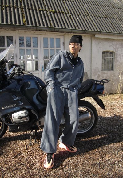 Hombre con chándal gris y zapatos rosas apoyado en una motocicleta negra frente a un edificio rústico con una gran puerta con ventanas.
