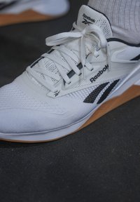 Tênis atléticos brancos com um cabedal em malha texturizada, detalhes pretos, atacadores robustos e sola de borracha gum, apresentando o logotipo da Reebok na lateral.