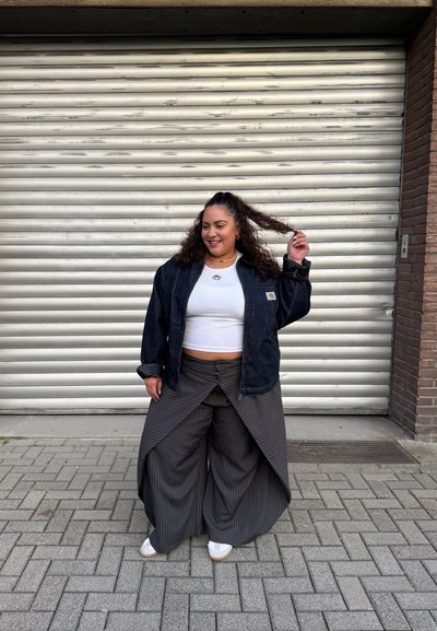 Mujer con cabello rizado que lleva un top corto blanco, chaqueta oscura, pantalones anchos de rayas finas y zapatos blancos, de pie en una calle pavimentada frente a una persiana metálica.