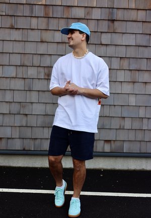 Hombre con gorra azul, camiseta blanca de gran tamaño, pantalones cortos azul marino y zapatillas azul claro, de pie sobre asfalto con una pared de tejas de madera de fondo.