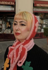 Sciarpa rosa e rossa lavorata a maglia con balze, abbinata a un maglione nero con applicazioni di orsetti colorati. Capelli morbidi con tonalità pastello.