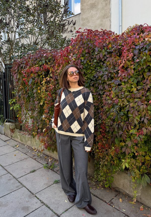 Una mujer con gafas de sol, un jersey de rombos y pantalones de pernera ancha está de pie en una acera junto a una pared cubierta de hojas rojas de otoño.