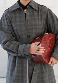 Trench-coat à carreaux gris avec de grands boutons, porté avec un sac à main rouge texturé embossé crocodile rond, et plusieurs bagues en argent à la main.