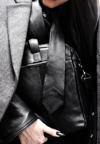 Sac en cuir noir avec fermeture éclair et bandoulière réglable, tenu sous un manteau gris. Détail notable d'un nœud sur le devant du sac. Texture lisse.