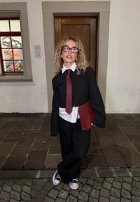 Czarny garnitur z luźną marynarką, biała koszula i bordowy krawat; nosi teksturowaną bordową torbę; kręcone włosy; stoi na brukowanej ulicy.