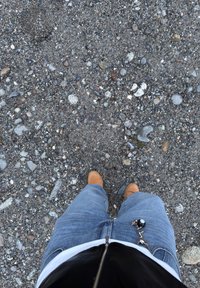 Bottes brunes sur un jean bleu, reposant sur une surface gravillonnée avec diverses petites pierres et cailloux, texturée de taches de terre plus foncées.