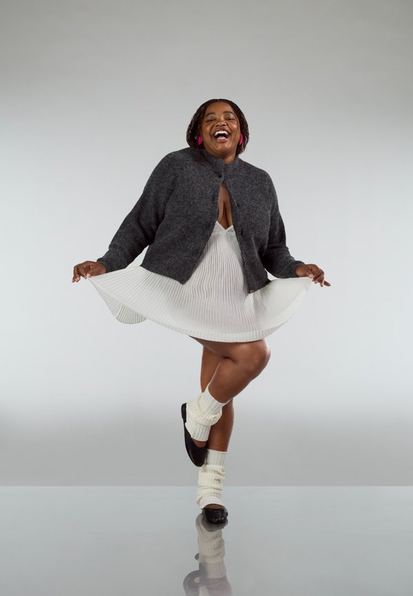 Grijze wollen cardigan over een witte geplooide jurk, gecombineerd met ivoorkleurige kniehoge sokken en zwarte ballerina's, op een reflecterend oppervlak.