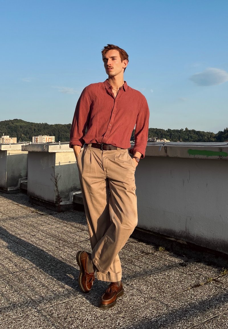 Jeune homme portant une chemise couleur rouille et un pantalon beige, debout, les mains dans les poches sur un toit sous un ciel bleu clair.