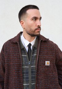 Veste à carreaux marron et orange avec une fermeture éclair, superposée à un pull gris à motifs, associée à une chemise blanche et une cravate noire.