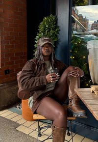 Άτομο που φοράει καφέ σακάκι, καπέλο και μπότες κάθεται σε τραπέζι εξωτερικού καφέ κρατώντας ένα φλιτζάνι καφέ, με φόντο φυλλώματα και τούβλινο τοίχο.