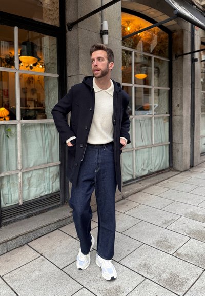 Abrigo azul marino con botones sobre un suéter color crema, combinado con pantalones de mezclilla oscuros y zapatillas blancas. Entorno urbano con grandes ventanas al fondo.