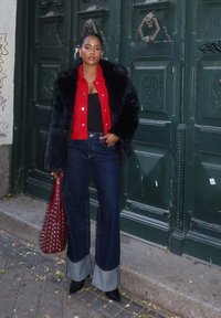 Palton din blană artificială neagră peste o jachetă roșie cu nasturi, un top negru, pantaloni din denim evazați cu manșete gri și o geantă roșie cu ținte. Stând împotriva unei uși verzi.